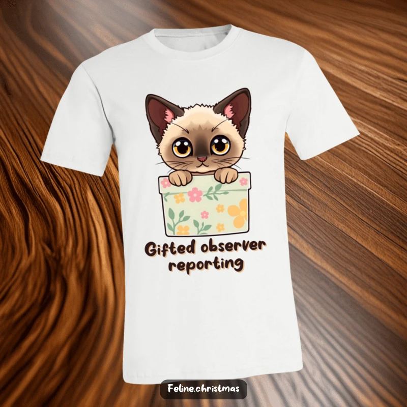 Funny t-shirt depicting a Siamese cat peeking out from a decorated gift box with wide eyes, full of holiday wonder.